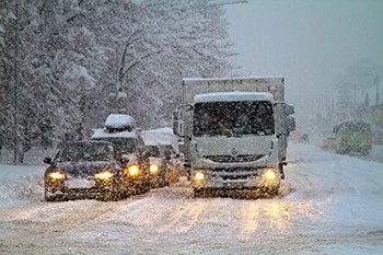 Photo of cars and trucks driving on a snow-packed road while more snow falls.
