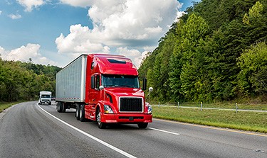 Photo of a red semi-truck driving on a highway surrounded by forest.