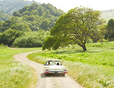 Photo of a couple in a silver convertible driving down a dirt road in the countryside.