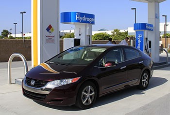 A photo of a dark maroon-colored sedan parked in front of a hydrogen fueling station.