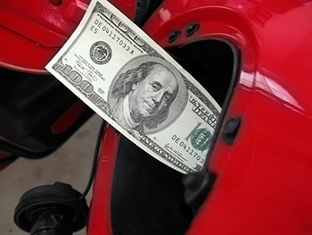 Photo of a one-hundred dollar bill sticking out of a fuel receptacle on a red car.