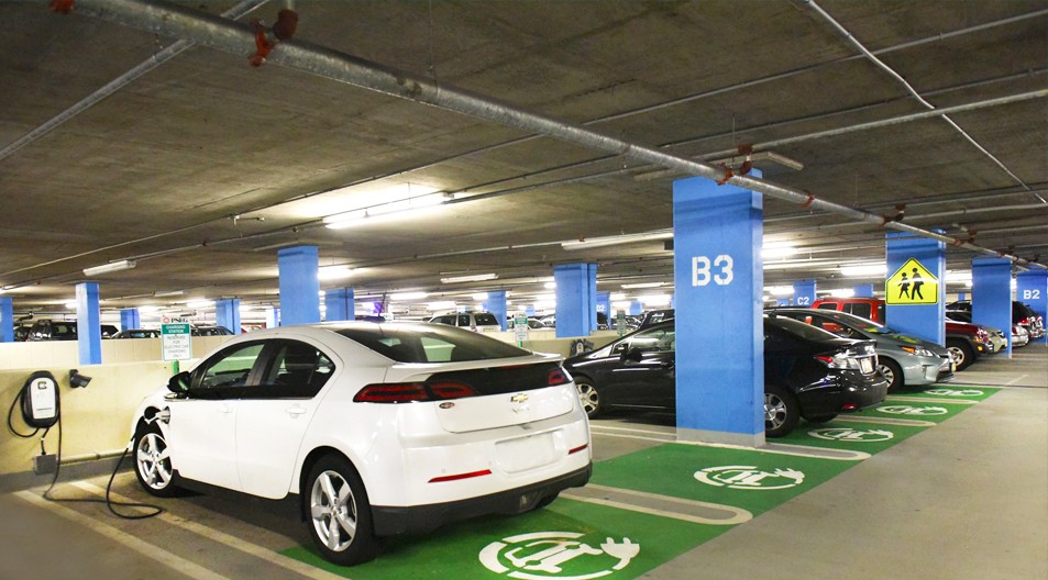 A photo of a white EV sedan plugged in to a charging station located inside of an employee parking garage.